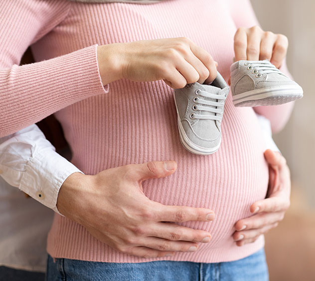 Die Schwangerschaft. Unkenntliches Paar wartet auf Baby, Ehemann hält winzige Schuhe in der Nähe des Bauches der schwangeren Frau posiert drinnen. Werdende Eltern, Familienmomente, Neues Lebenskonzept.